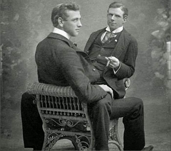 Two handsome Victorian men in formal suits sitting on an ornate bench in a vintage black and white photo.