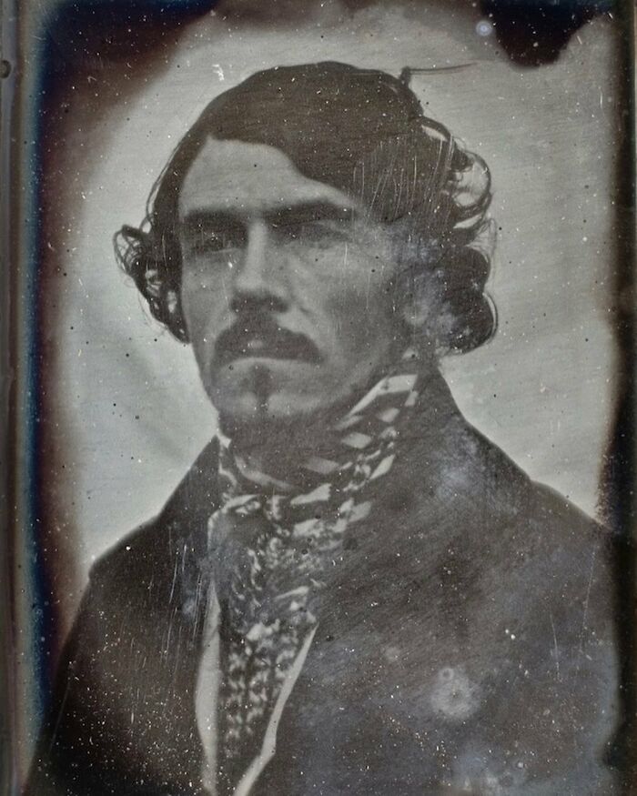 Victorian man with styled hair and mustache, wearing a patterned cravat and dark coat in a historic black and white photo.