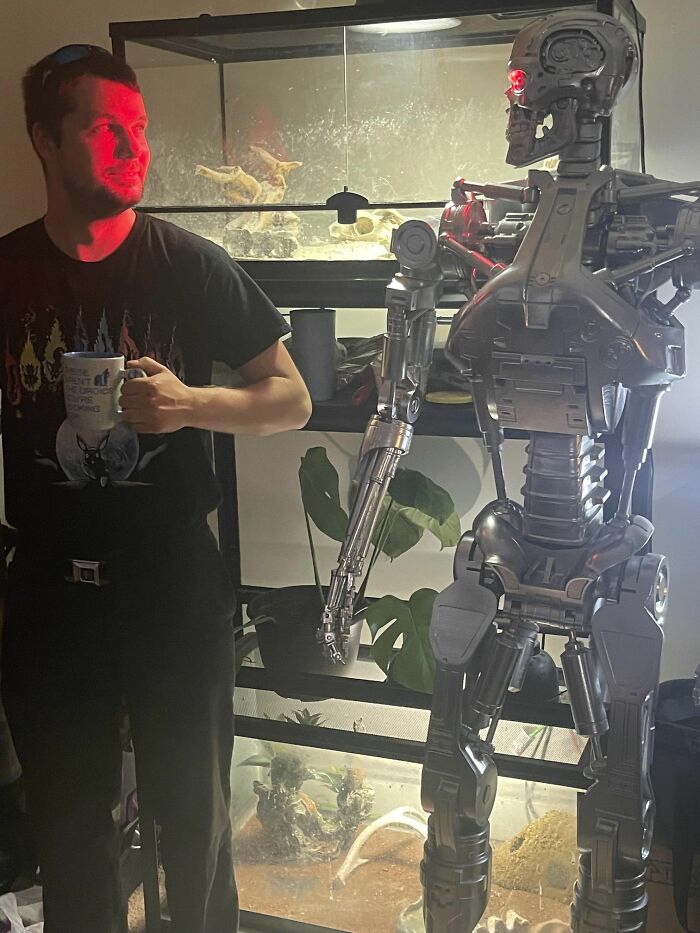 Man holding a mug looking excited next to a life-sized robot figure with glowing red eyes in a dimly lit room.