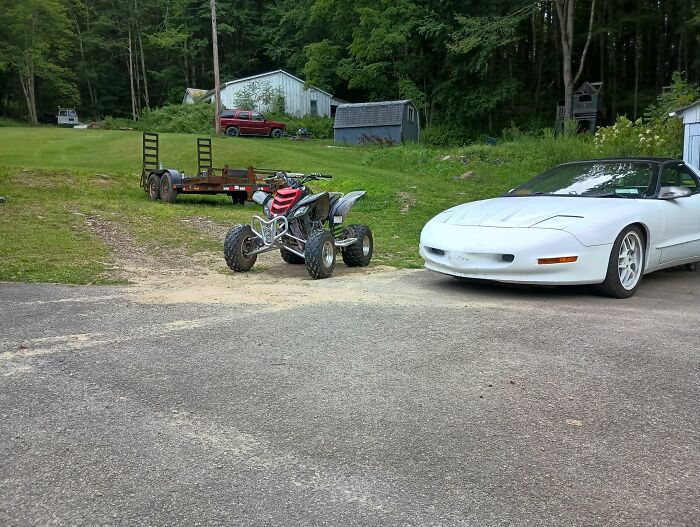 White sports car and red ATV parked on driveway near trailer and wooded area, a scene any man would be excited to see