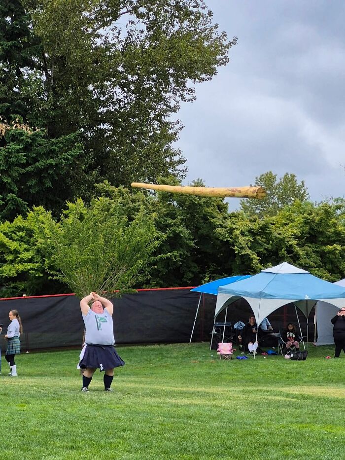 Man wearing a kilt throwing a large wooden log at an outdoor event, captured in mid-air on a grassy field.
