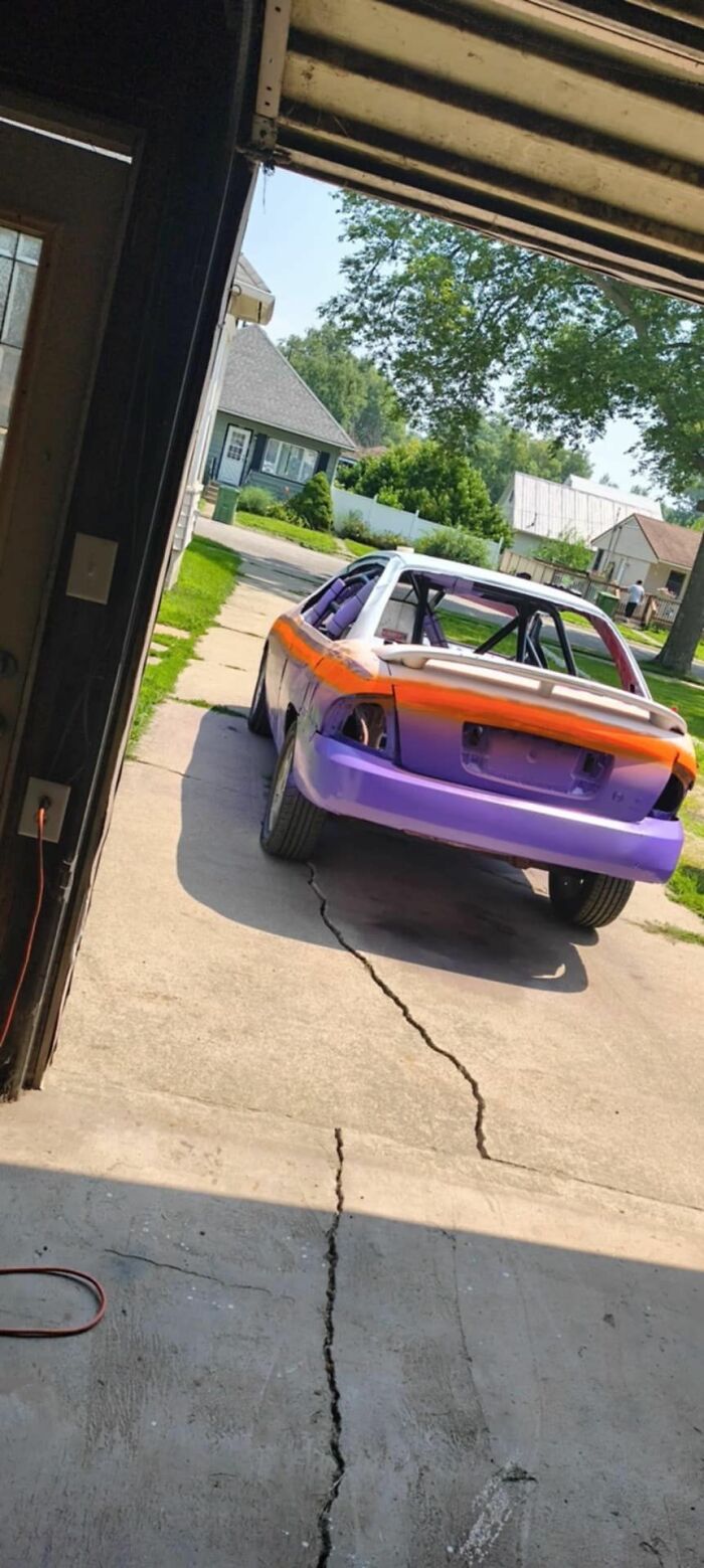 Purple and orange car body in driveway, viewed from garage with c*****d concrete, surrounded by suburban houses and trees.