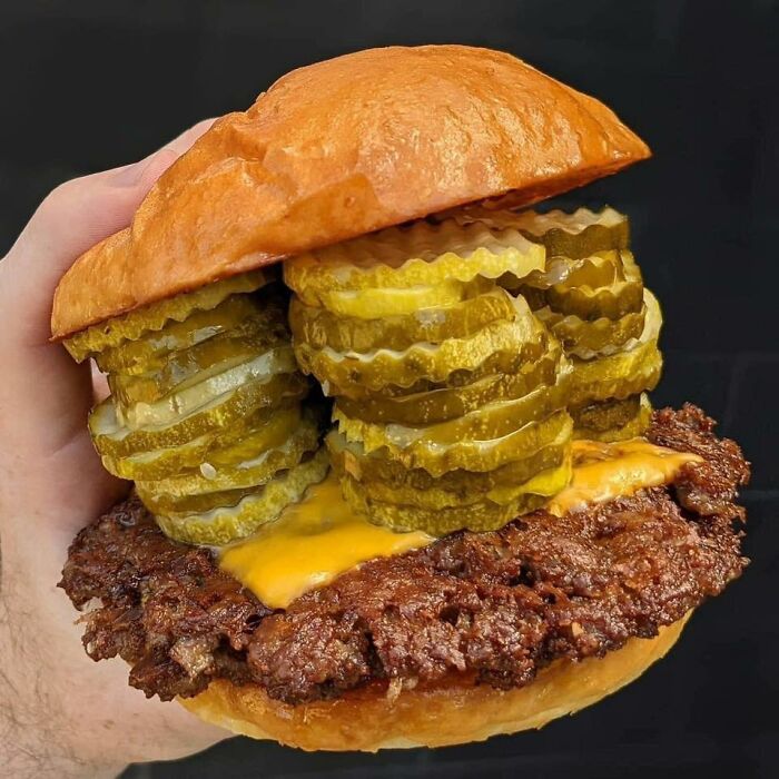 Hand holding a cheeseburger loaded with crispy beef patty and stacked pickles, a picture any man would be excited to see.
