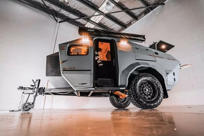 Off-road camper trailer with rugged tires and open door inside a modern industrial garage setting.
