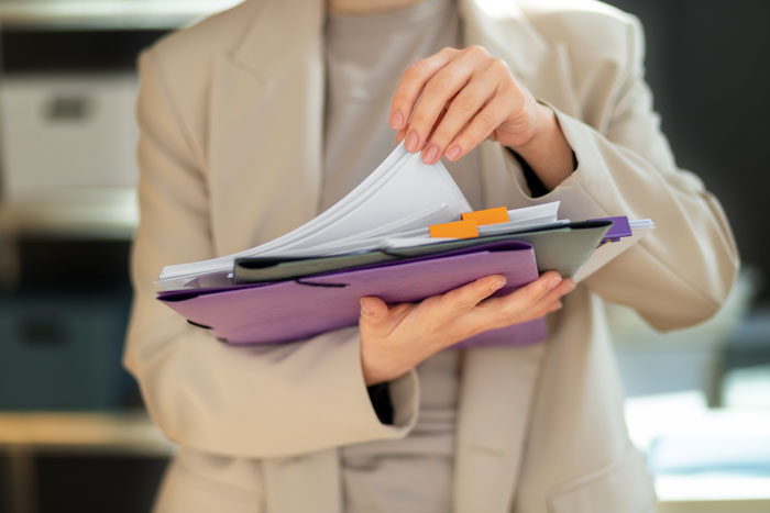 Person in beige blazer holding and flipping through a folder filled with documents on toxic newbie and HR issues.