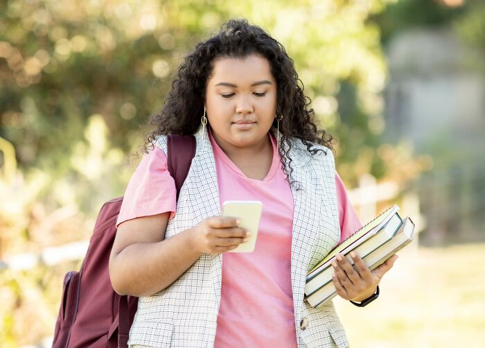 Young woman holding books and looking at her phone outdoors, depicting stories of people getting savagely rejected.