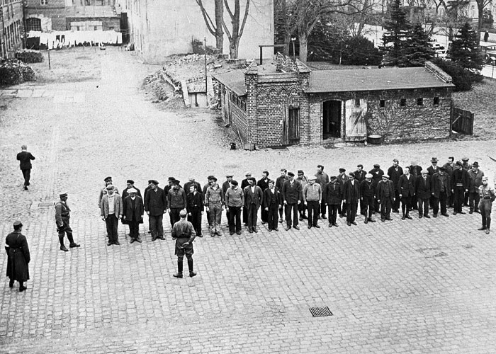 Black and white photo of a group of men standing in a line under watch during a historical event people survived.