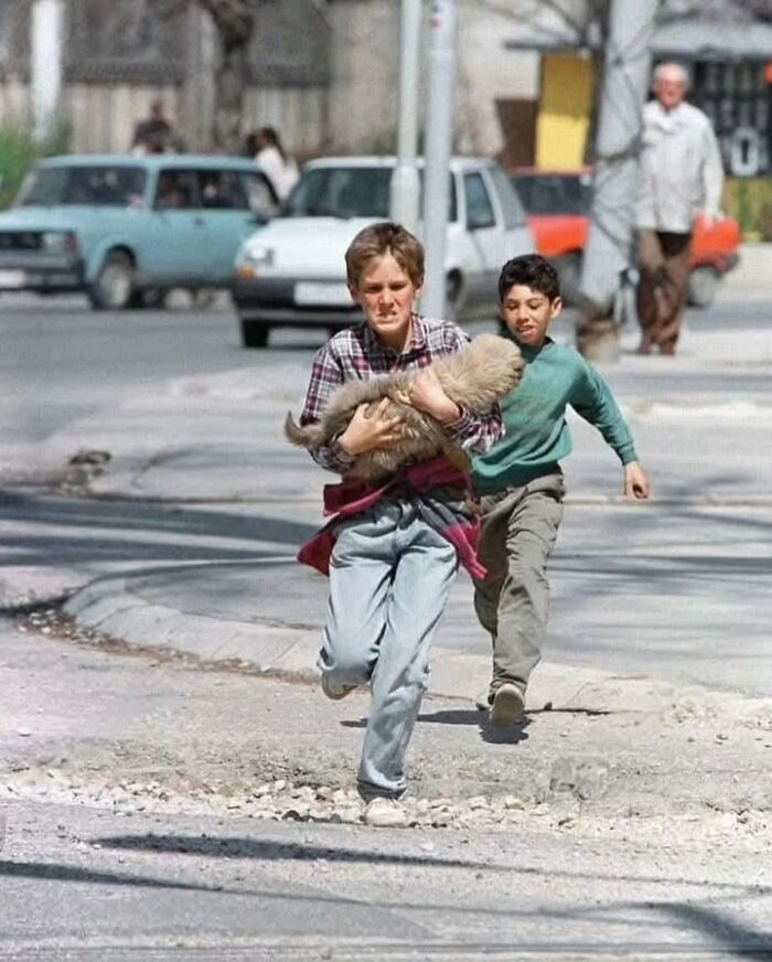 Two boys running on a street, one holding a fluffy pet, in a historical photo capturing pets and childhood moments.