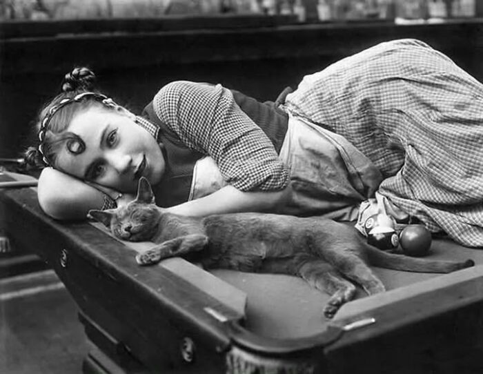 Black and white historical photo of a woman lying on a pool table with her pet cat, showcasing unique pet moments.