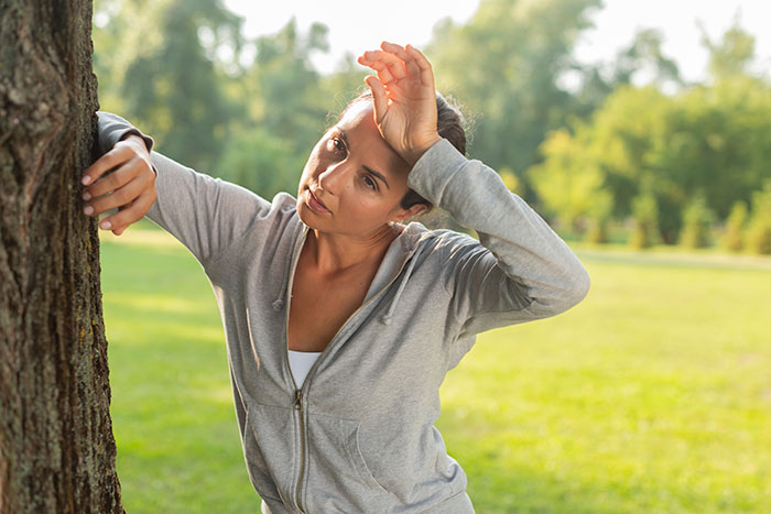 Woman in gray hoodie leaning on a tree and wiping sweat, showing surprising body quirks on a sunny day outdoors