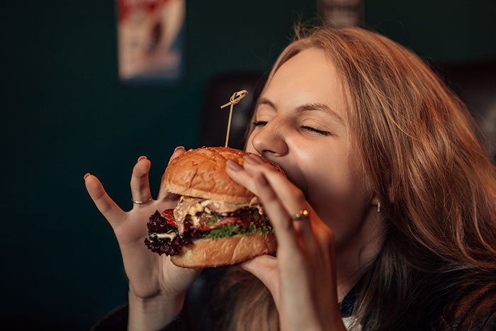 Young woman enjoying a large burger indoors, showcasing surprising body quirks related to eating and senses.