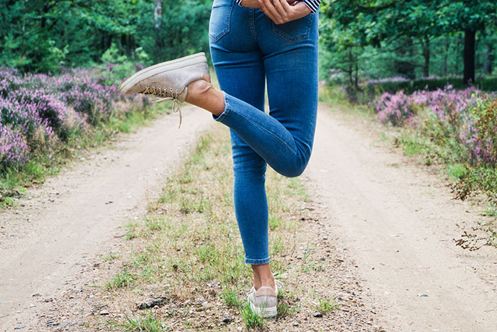 Person standing on a dirt path lifting one leg, wearing blue jeans and beige shoes, showing surprising body quirks outdoors.