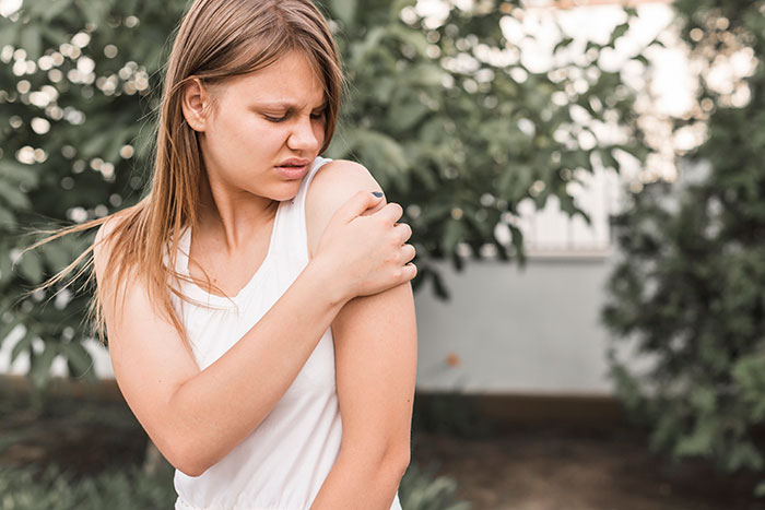 Young woman in a white tank top outdoors holding her shoulder in discomfort showing surprising body quirks and reactions.