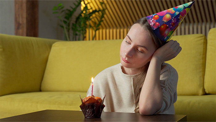 Woman wearing a birthday hat looks disappointed sitting alone with a cupcake candle, hinting at husband’s birthday surprise gone wrong. - 1