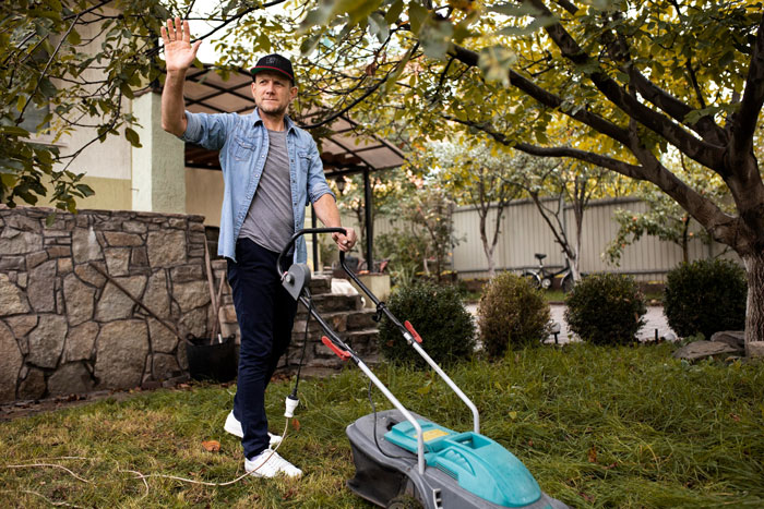 Man mowing lawn wearing casual clothes and cap, waving hand outdoors near stone wall and trees on overgrown grass Man mowing lawn wearing casual clothes and cap, waving hand outdoors near stone wall and trees on overgrown grass