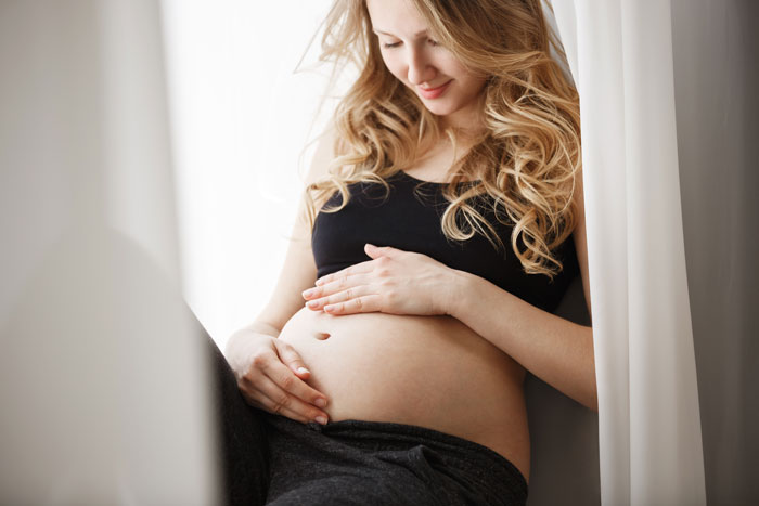 Pregnant woman smiling gently while sitting by the window, holding her belly and embracing her pregnancy moment. - 5