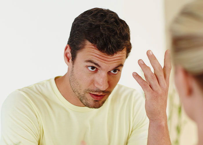 Man with beard and short hair wearing a yellow shirt, expressing frustration while talking to a woman, relationship conflict theme.