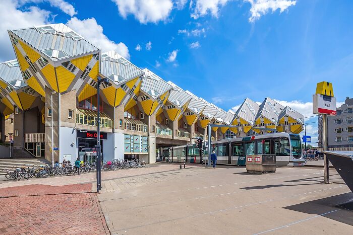 Cube-shaped iconic architectural homes in an urban setting with a tram and bicycle parking under a bright blue sky.
