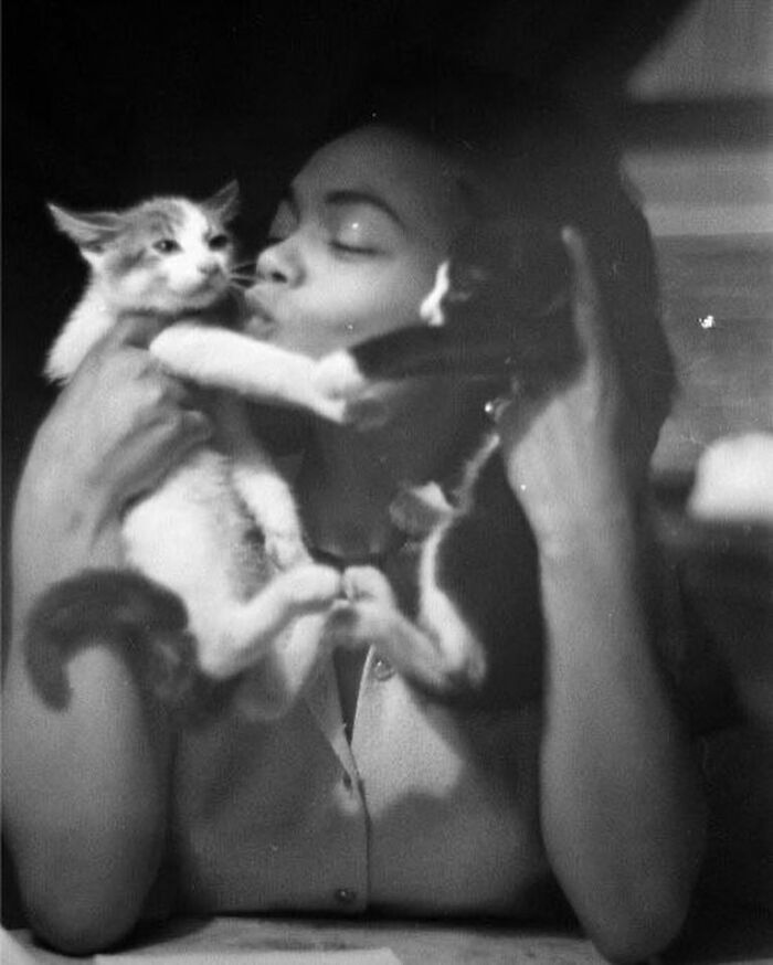 Black and white photo of a woman holding and kissing two cats, a weird history pic showing an unusual affectionate moment.