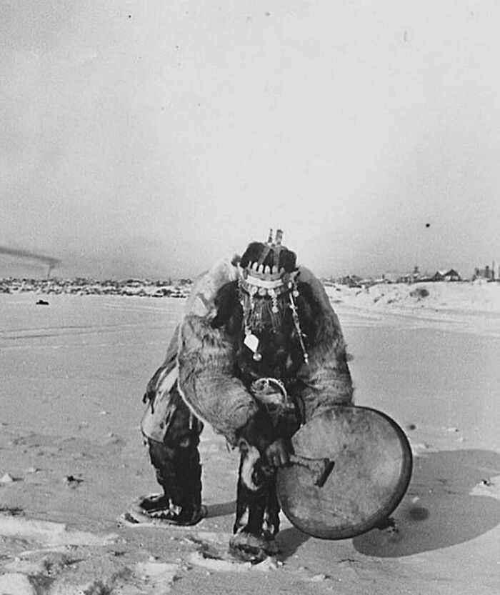 Indigenous shaman dressed in traditional attire holding a drum on snowy terrain in a weird history pic from the past.