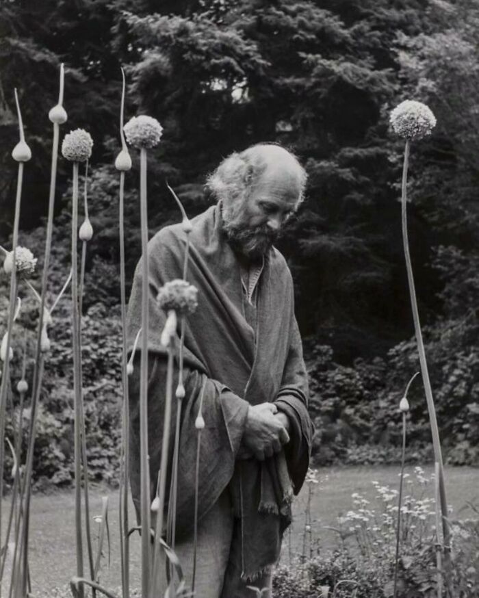 Black and white photo of a man in a robe standing thoughtfully among tall plants in a weird history image.