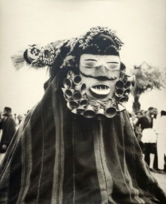 Black and white photo of a person wearing an unusual traditional mask at a weird history event outdoors.