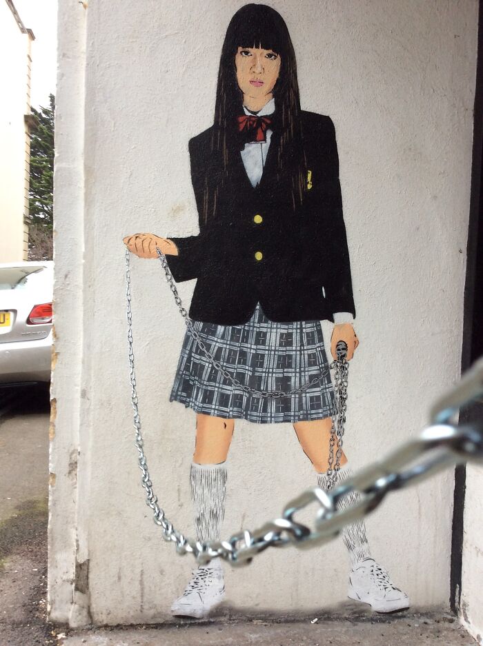 Street art mural of a girl in a school uniform holding a chain, blending real and painted elements on an urban wall.