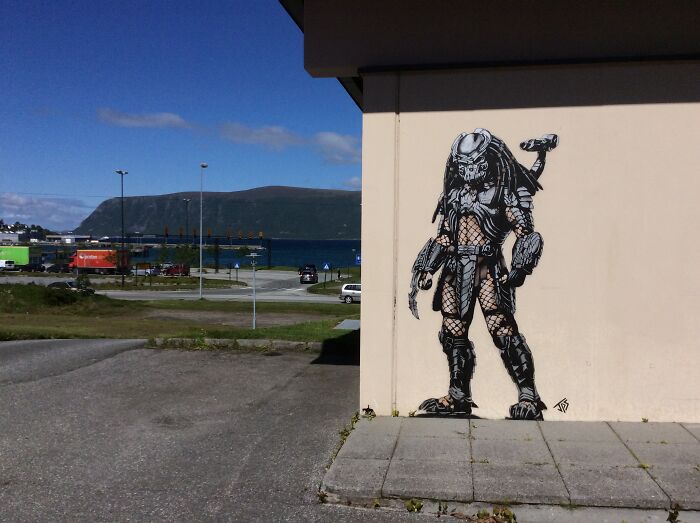 Street art mural of a Predator character painted on a building wall near a coastal area with clear sky.