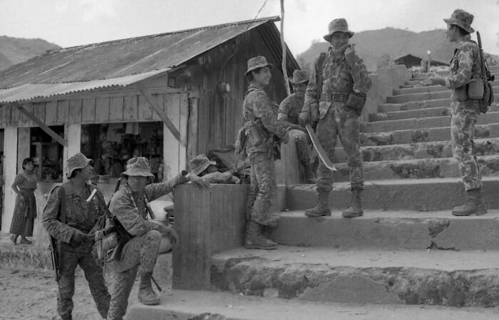 Black and white photo of soldiers in camouflage uniforms gathered near a rustic building and stone steps in a weird history scene.