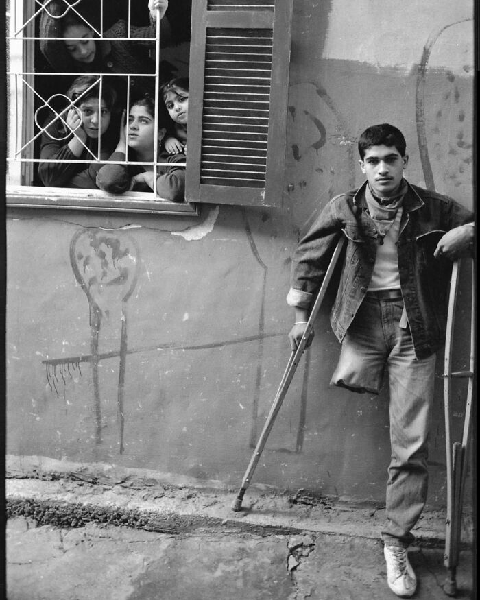 Young man with crutches and missing leg standing by a wall while four children watch from a barred window in a weird history pic.