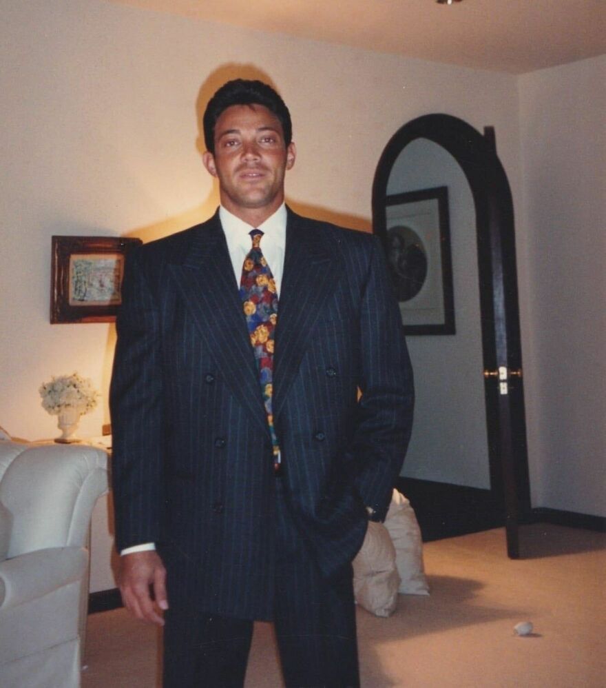 Young Jordan Belfort in a dark pinstripe suit and floral tie standing in a living room, showcasing his former millionaire lifestyle.