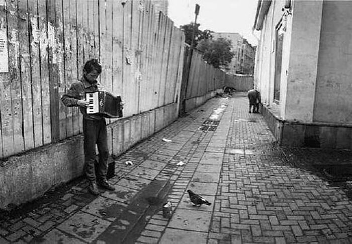 Boy playing accordion on a narrow street with pigeons in a black and white weird history photo.