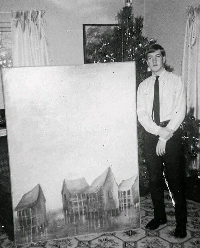 Black and white photo of a young man standing next to a large painting in a vintage home setting, weird history pics.
