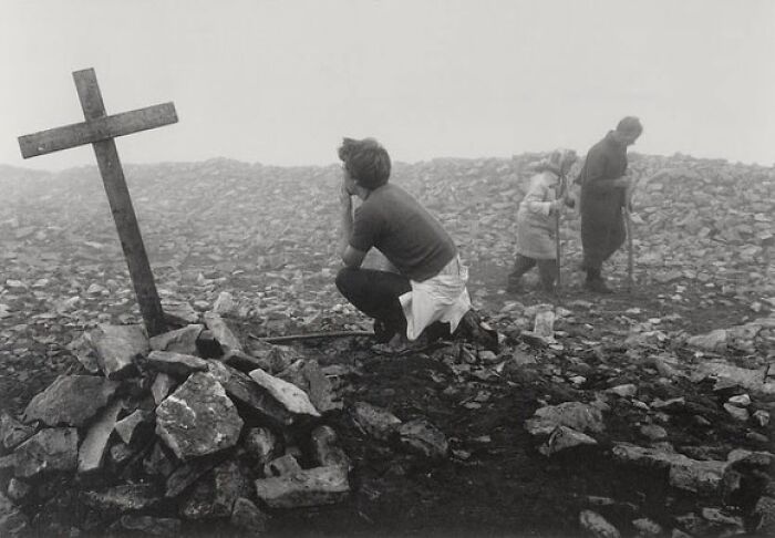 Woman kneeling by a grave with a wooden cross in a rocky, foggy landscape in a weird history pic from the past.