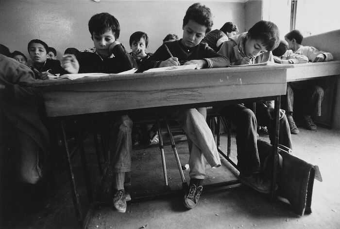 Black and white photo of children writing at a wooden desk in a classroom capturing a weird history moment from the past.