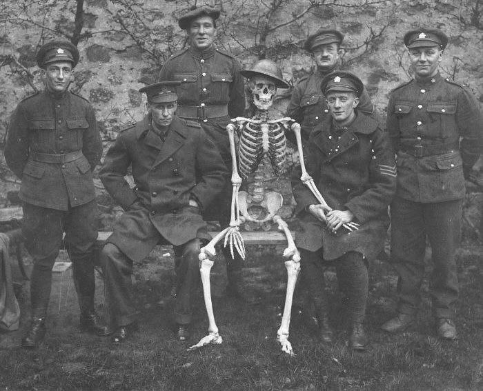 Group of World War I soldiers posing with a skeleton wearing a helmet in a weird history photo from the past.
