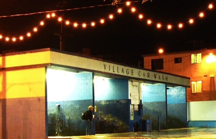Man with backpack standing at a lit Village Car Wash at night, capturing everyday life in a street photo scene.