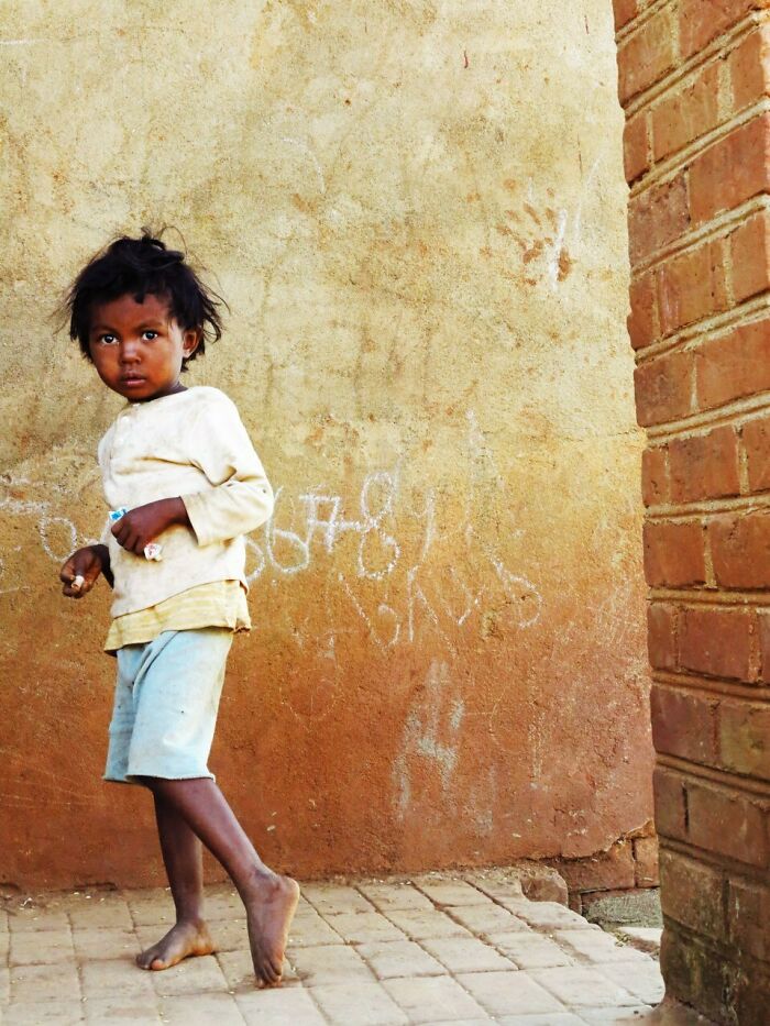 Child standing barefoot near a textured wall with chalk markings, captured in a street moment by Stéphane Kyndt.