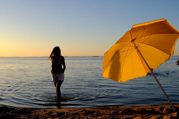 Silhouette of a woman walking in shallow water at sunset beside a bright yellow umbrella in street photos of everyday life.