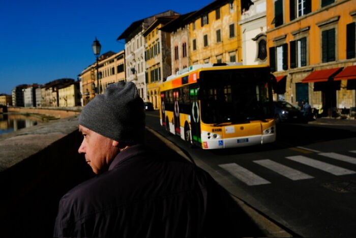 Man wearing a gray beanie walking along a street near a colorful bus with historic buildings in bright sunlight.