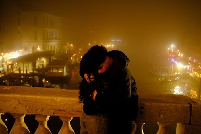 Couple embracing at night on a bridge, warm street lights glowing in fog, a candid street photo showing everyday life.