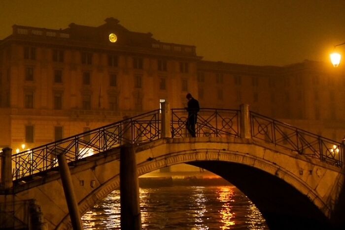 Person standing on an illuminated bridge over water at night in a street photo capturing everyday life.