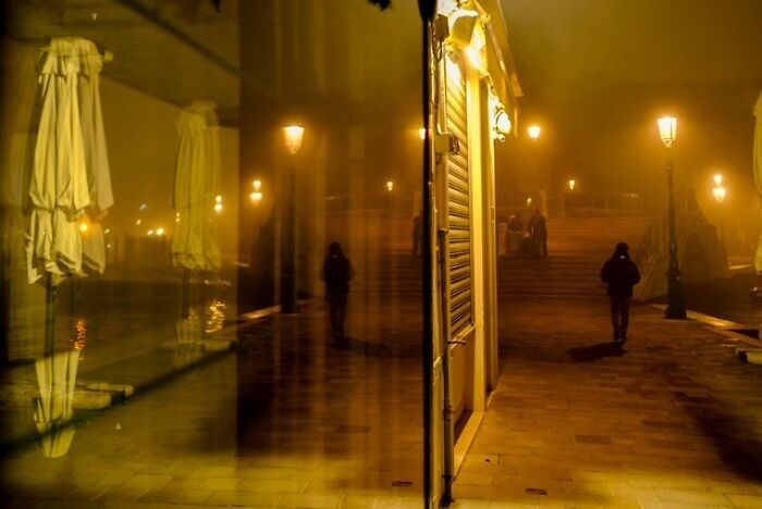 Street photo capturing a solitary figure walking at night with warm street lamps and reflections in a glass window.