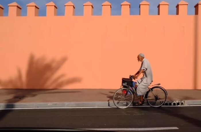 Elderly man riding a bicycle along a street with a peach-colored wall and palm shadow in urban everyday life scene.