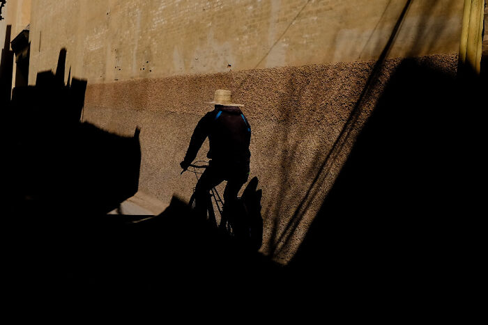 Silhouette of a person riding a bicycle along a textured wall in a street photo capturing everyday life moments.