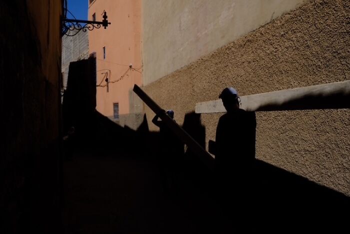 Shadowed figures carrying wood beams along a narrow street in a street photo capturing everyday life.