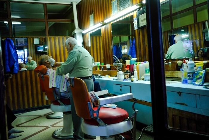 Barber trimming beard of elderly man in a vintage barbershop, a candid street photo capturing everyday life moments.