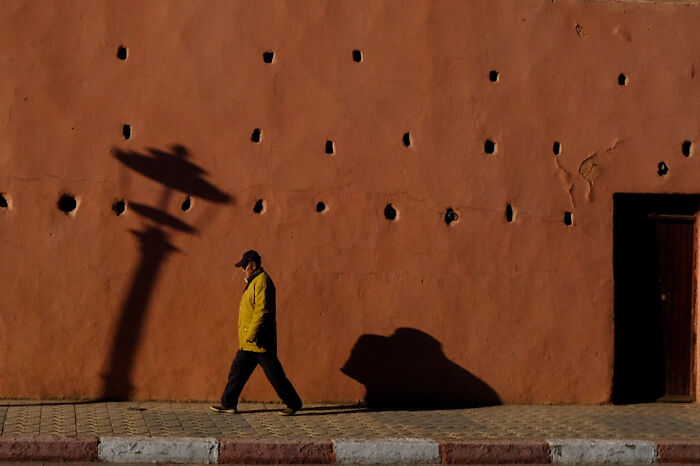 Man walking along a textured wall with shadows creating abstract shapes in a street photo capturing everyday life moments.
