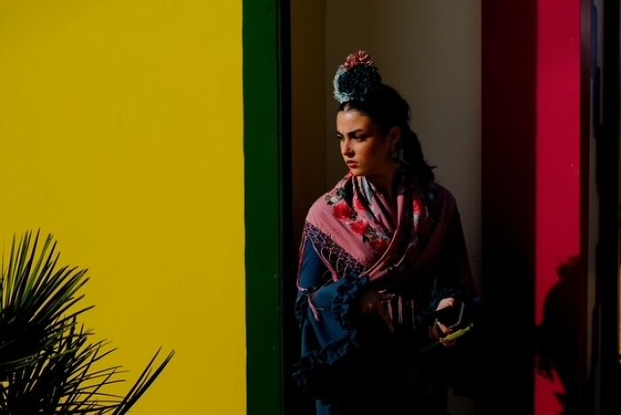 Young woman wrapped in a colorful shawl standing against vibrant yellow and red walls in a street photo capturing everyday life.