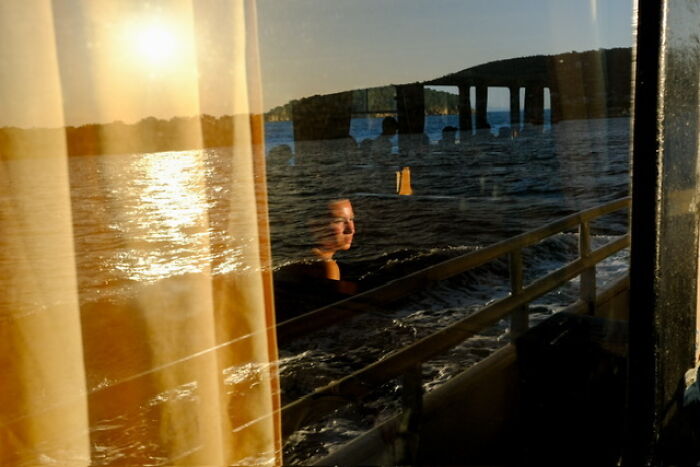 Reflection of a woman sitting by a window overlooking water at sunset, capturing everyday life in a street photo.